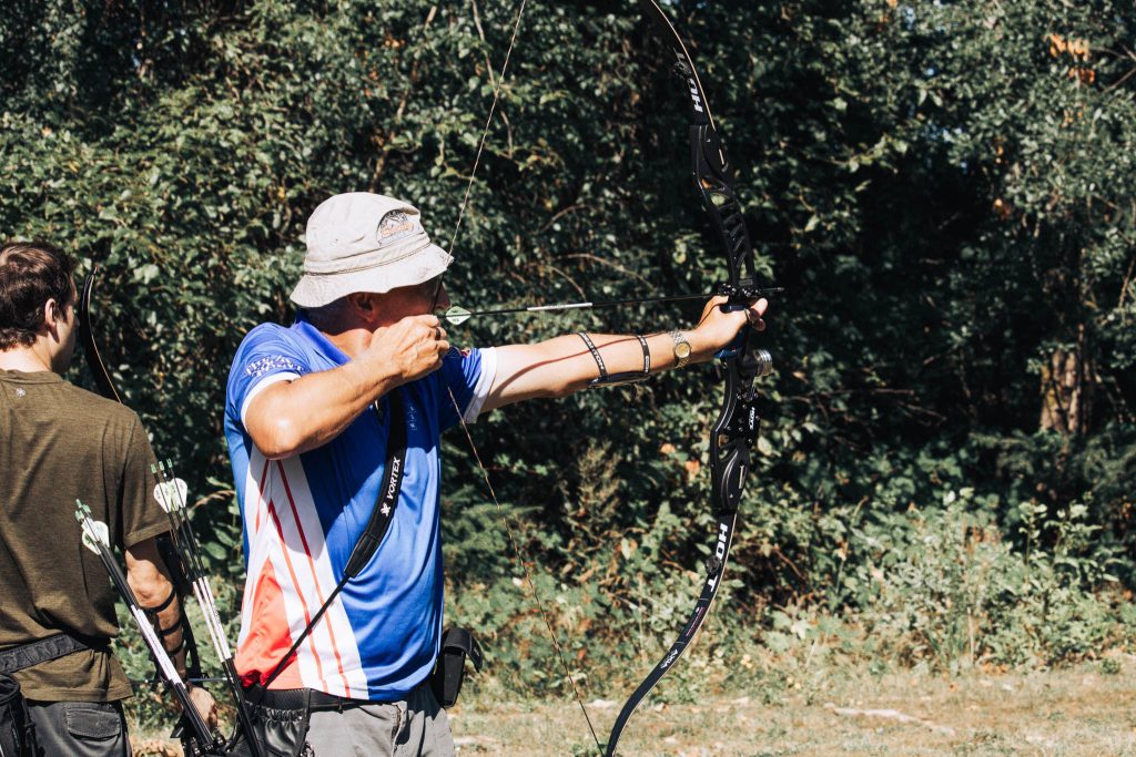 An elder man in a bucket wearing a royal blue jersey that has red and white strips on the right side. He is shooting his barebow outside on a FITA field